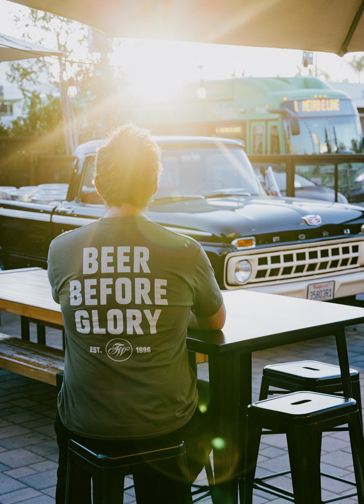 Man wearing a military green Firestone Walker "Beer Before Glory" t-shirt, back view, seated at a table with a vintage truck in the background, sunlit