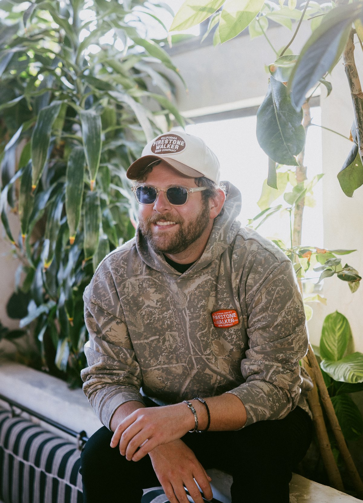 Smiling man in a Firestone Walker Trailhead Hoodie in woodland camo and a cream Firestone Walker cap