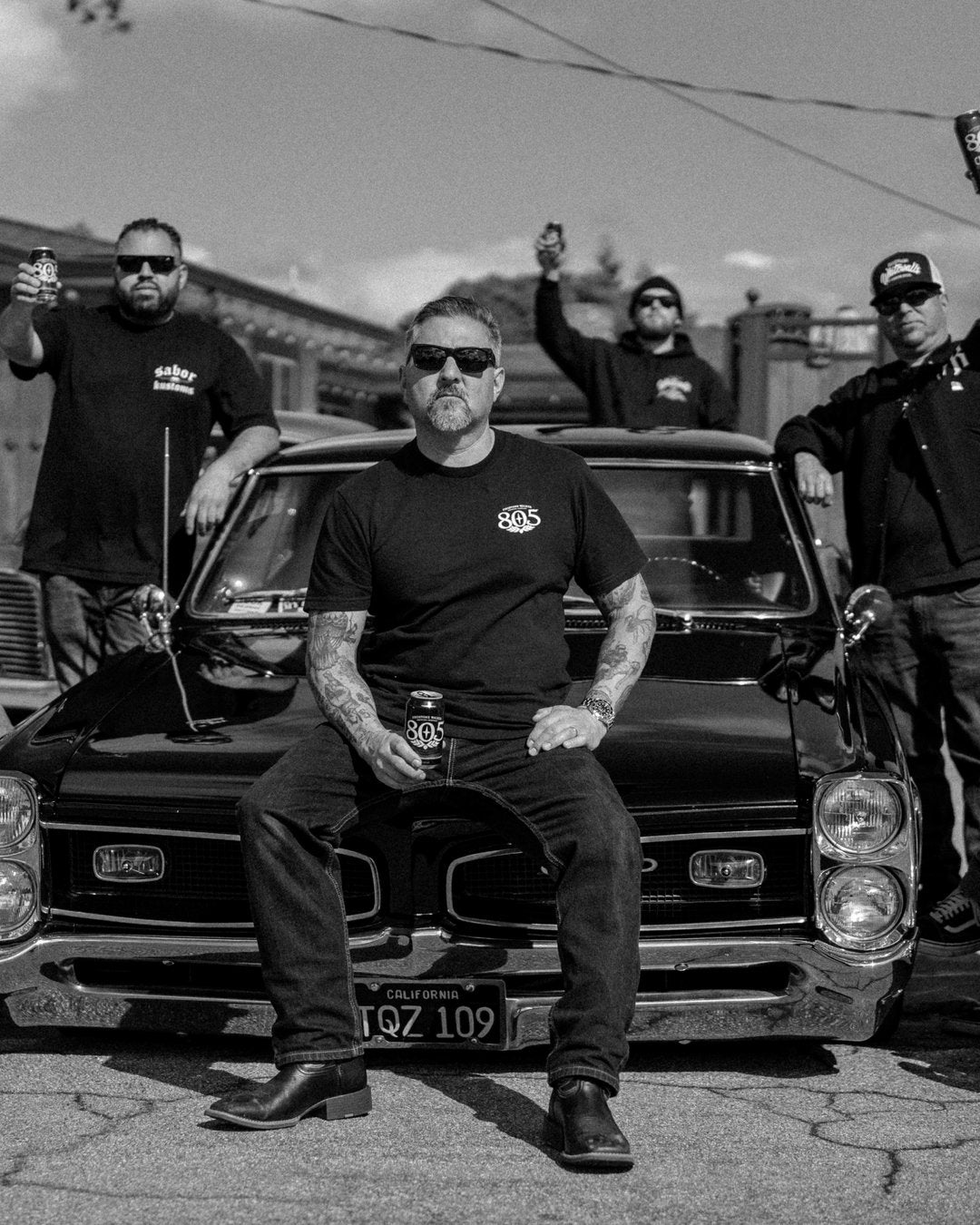 Black-and-white photo of four Sabor Kustoms members gathered around a classic black car, one seated on the bumper holding an 805 can while the others raise their cans behind him