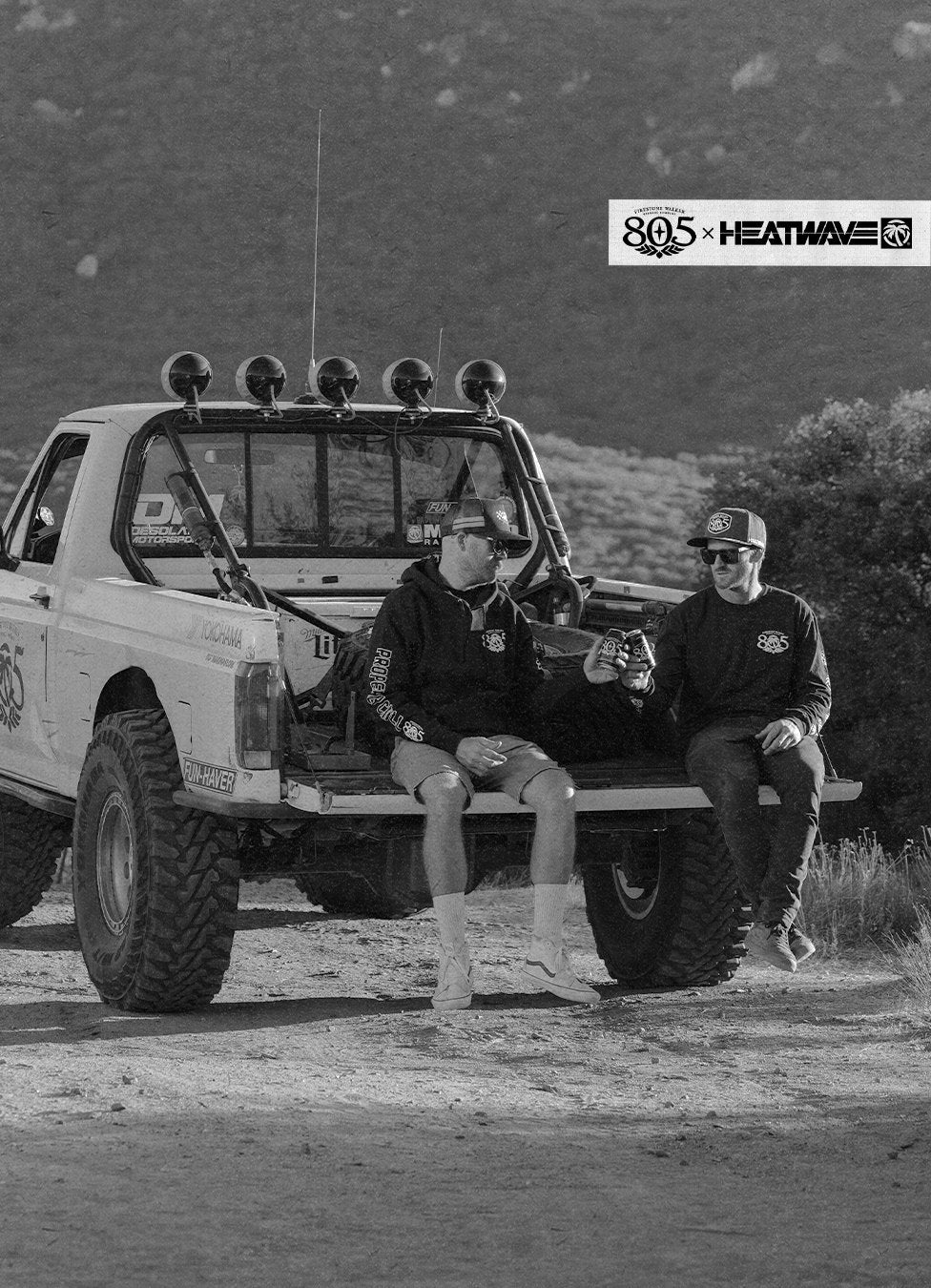 Black-and-white photo of two friends in 805 x Heat Wave hoodies, hats and sunglasses sitting on the tailgate of an off-road race truck, clinking 805 cans, with the 805 x Heat Wave logo in the corner
