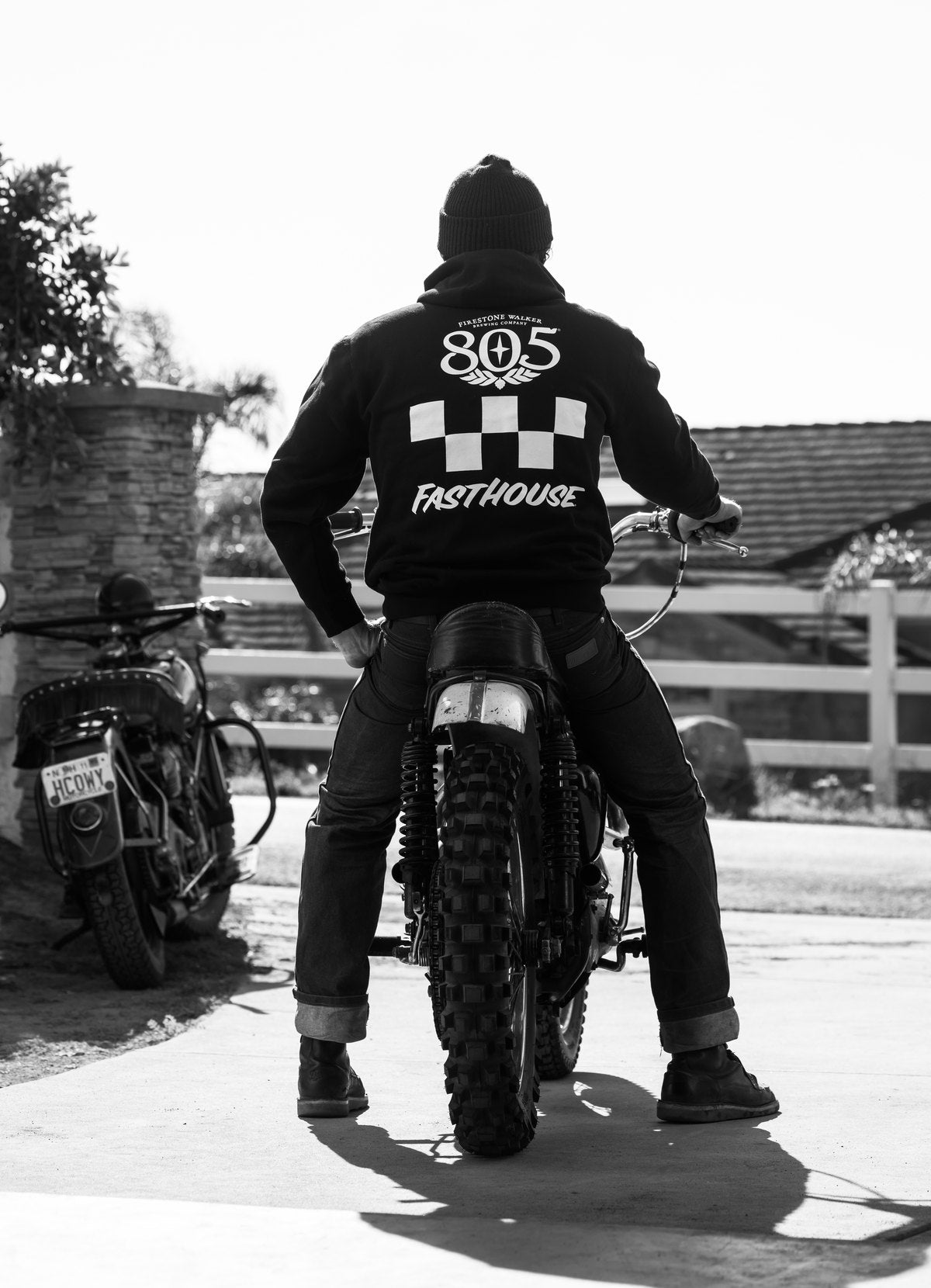 Black-and-white photo of a rider from behind on a vintage dirt bike, wearing a black 805 x Fasthouse hoodie with the 805 logo, checkered flag and Fasthouse wordmark on the back