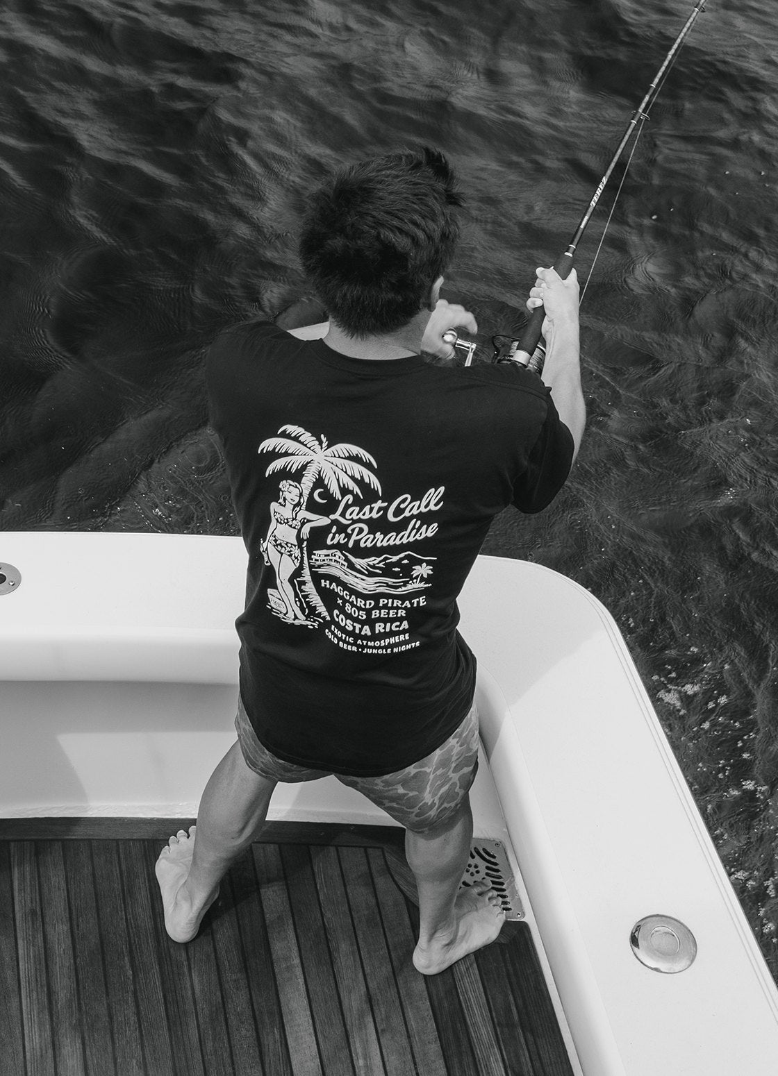 Black-and-white overhead photo of a man reeling in a fish off the back of a boat, wearing a black 805 x Haggard Pirate "Last Call in Paradise" Costa Rica tee
