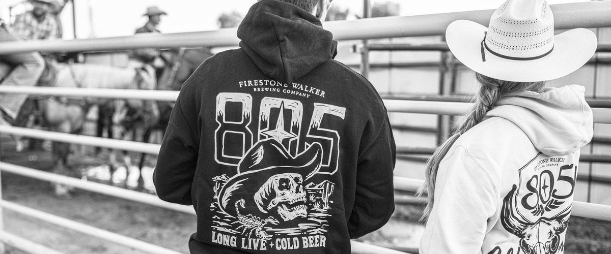 Two people at a rodeo pen seen from behind, wearing a black 805 hoodie with a skull-in-cowboy-hat graphic and "Long Live Cold Beer," and a white 805 Central Coast hoodie with a bull skull graphic, with horses and riders behind the fence