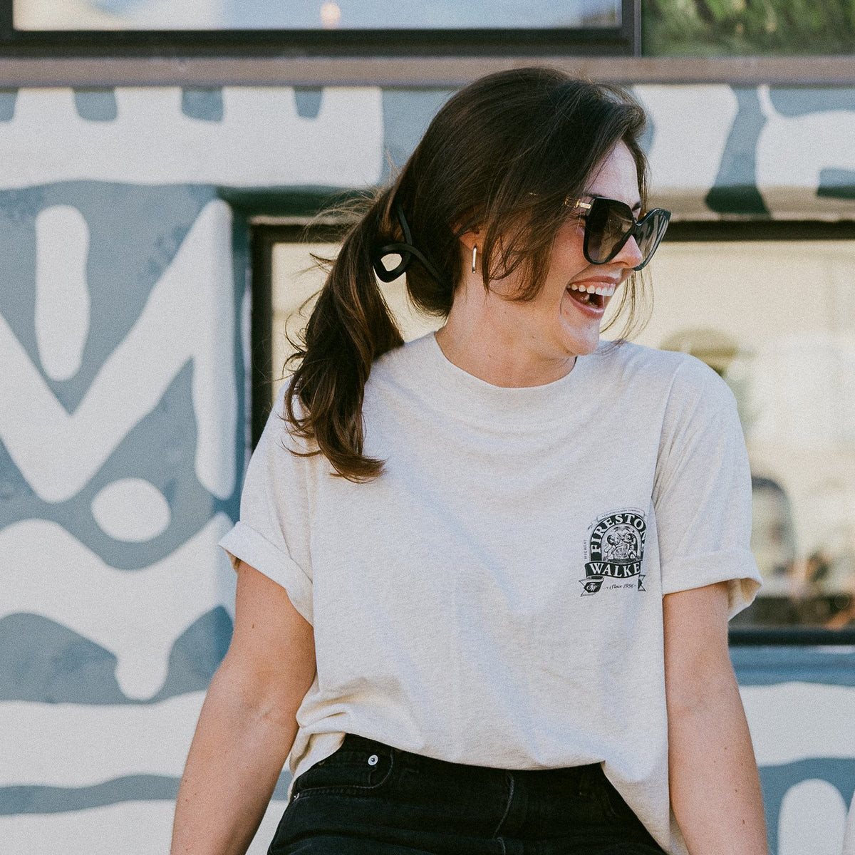 Woman with a dark ponytail and sunglasses laughing against a painted mural wall, wearing a cream Firestone Walker tee with a small logo on the chest